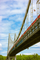 A modern pedestrian iron cable-stayed bridge with supporting structure and a lantern against a blue cloudy sky in summer day. View from below on a high red bridge in Kyiv, Ukraine. Urban architecture.
