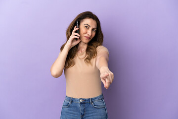 Middle aged caucasian woman using mobile phone isolated on purple background pointing front with happy expression