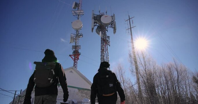 An aesthetic videography of a couple of documentary makers walking toward cellular communication towers, as it snows mildly in the sun