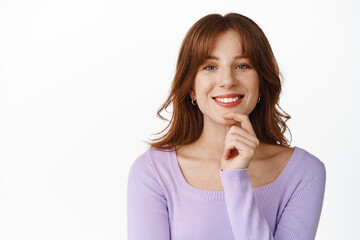 Smiling modern girl with red lips and make up, looking successful and happy, touch chin, have interesting idea, listening with friendly face, standing against white background