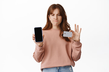 Finance and money. Frowning and disappointed redhead girl shows mobile phone screen and credit...