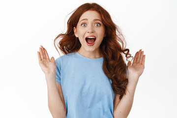 Portrait of excited redhead girl screaming from amazement and joy, raising hands up and look surprised astonished, standing against white background
