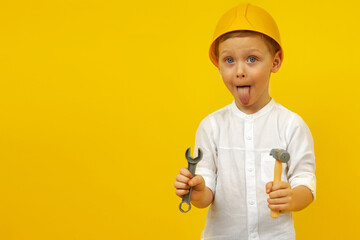 A cute boy in a hard hat, with a wrench and a hammer in his hands poses on a yellow background showing his tongue.