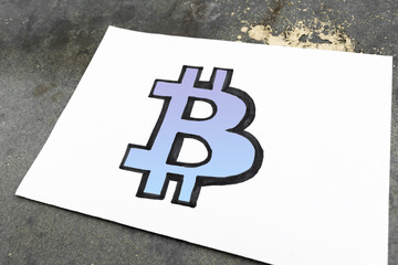 blue Bitcoin Sign Drawn on the sheet of White Paper on the gray background. The concept of investments in digital assets.
