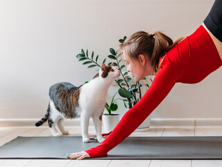 Girl in red sportswear with cute funny cat practicing yoga at home. Indoor and outdoor workout....