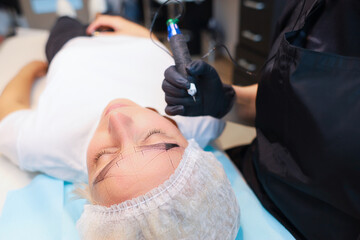 The client lies under the permanent eyebrow makeup procedure with a drawn sketch on his face, and...