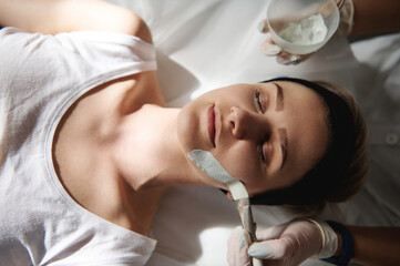 Top view of woman receiving spa beauty treatments and beautician applying alginate mask on her face. Spa professional skin care concept