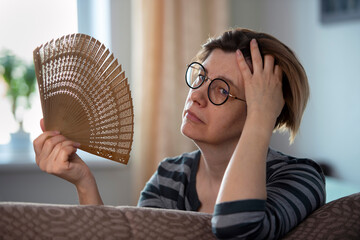 Exhausted middle aged woman waving her fan, suffering from menopausal symptoms, experiencing hot...