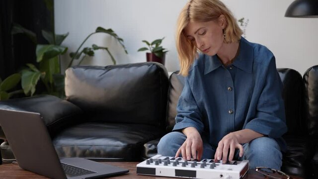 Young woman playing on midi controller in living room with couch, producer of music with laptop on table. Student at online music school, young producer
