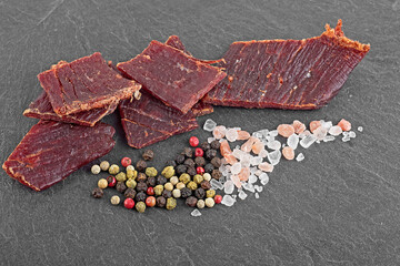 Beef jerky pieces, peppercorns and sea salt on black stone table. Dried peppered beef jerky.