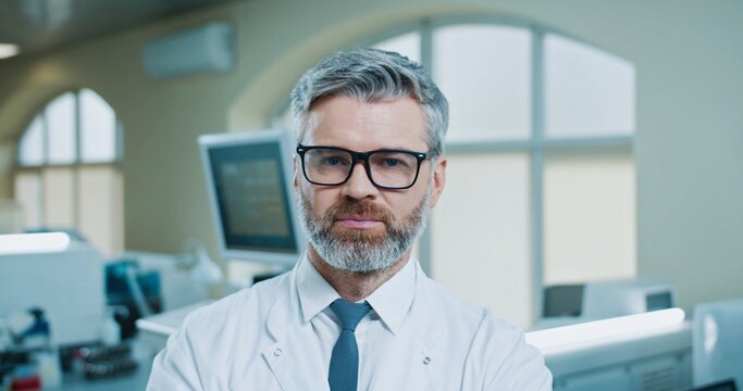 Close up portrait of cheerful Caucasian bearded senior male professional medic standing in clinic laboratory looking at camera and smiling. Handsome happy worker at workplace in hospital lab
