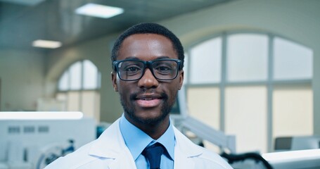 Naklejka na ściany i meble Close up of African American joyful young handsome male doctor in glasses looking at camera and smiling at workplace in hospital lab. Medical worker, laboratory specialist, medicine concept