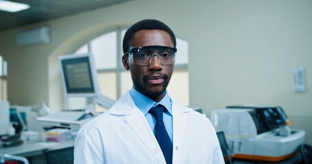 Fototapeta premium Portrait of happy African American young handsome male physician in goggles looking at camera and smiling standing at work in hospital lab. Medical profession, laboratory specialist, medicine concept