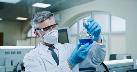 Close up of middle-aged Caucasian man pharmacist in goggles and gloves holding glassware with blue liquid in modern laboratory. Male medical scientist works on coronavirus cure. Chemical research