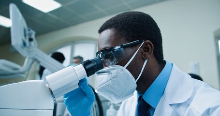 Close up of young African American busy male medical scientist working in laboratory on virus cure. Man specialist in mask and goggles looking at blood cells through microscope modern equipment