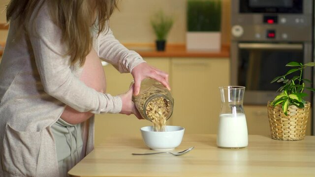 Beautiful pregnant woman preparing healthy breakfast. Delicious food during pregnancy made muesli and milk. Mom put cereal in bowl and pours it over. Stir food with spoon. Close up pregnant belly.