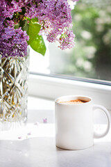 A bouquet of flowers on the window and coffee in a white cup in the rays of sunlight. A bouquet of lilacs and a cup of coffee.