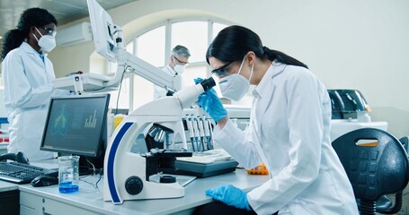 Side view of young Caucasian female medical scientist in mask and gloves sitting at laboratory using modern technology looking in microscope conducting experiment. Lab assistant making analysis