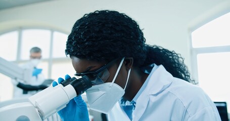 Close up of busy African American ?????? medical researcher in white coat sitting at hospital laboratory using microscope, scientist looking at blood cells through equipment, experiment concept