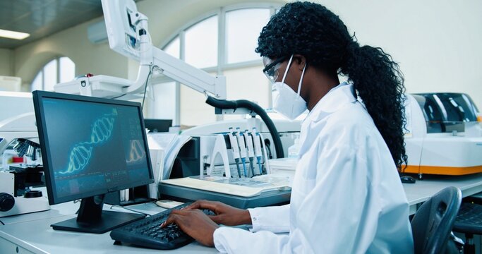 Side view of African American young female medical expert sitting at workplace in lab working typing on computer looking at molecular structure. Scientist using computer in his modern laboratory