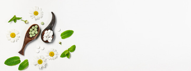 Concept of herbal medicine. Natural medical tablets on the wooden spoons with chamomile flowers and mint leaves on white background long banner format. Alternative medicine and health care.