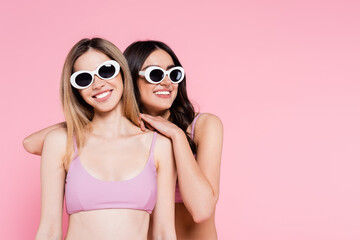 Smiling woman in sunglasses hugging friend isolated on pink.