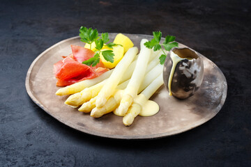 Traditional steamed white asparagus with cured ham and boiled potatoes garnished with sauce hollandaise served as close-up on a rustic design plate