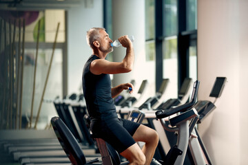 Mature sportsman having water break while exercising on stationary bike in a gym.