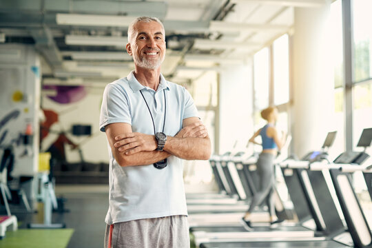 Happy mature personal trainer standing with arms crossed in a gym and looking at camera.