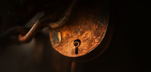 An old vintage heavy lock, covered in rust, hangs on a thick chain in the twilight, closing something. A mystery.