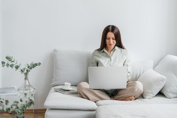 Fototapeta premium Portrait of a young happy female freelancer sitting on the couch and working on project, watching movie on laptop, studying, blogging, resting and chatting online