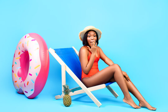 Oops. Shy black lady in bikini chilling in lounge chair, covering her mouth, making mistake on blue studio background