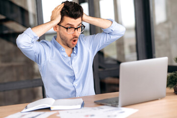 Shocked caucasian male manager, broker or freelancer, working in office, using laptop, incurring huge losses, overworked, stressed, looking at screen in surprise, holding his head with his hands