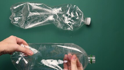 POV female hands crumple and crush transparent PET plastic bottle for recycling. Crumpled used bottles on green background. First point view preparation of recycled household trash as zero waste