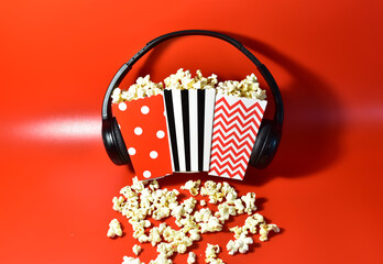 Popcorn in colored cardboard packaging with headphones for listening to music and watching a movie. Heap of salted popcorn in paper striped bucketon a red background.