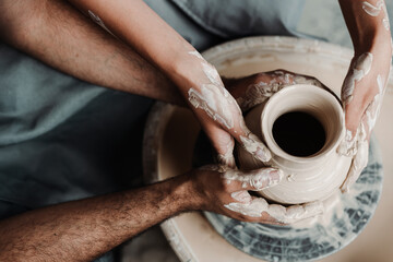Fototapeta na wymiar Top view, Close-up of the hands of an African American man and a white woman sitting together at a potter's wheel and making a clay pot. Romance, dating in the masseter's hobby