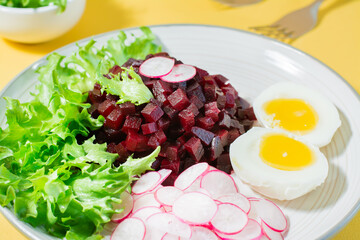 A dietary dish made from vegetables. Beet tartare, radish, frisee salad and boiled egg on a plate on a yellow background. Hard light. Close-up