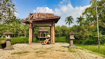 The guy sits in a Vietnamese hat near gazebo and the arch. Lake, trees and Asian architecture. Tropical landscape in Ao Dai Museum. Ho Chi Minh City (Saigon). Vietnam. South-East Asia