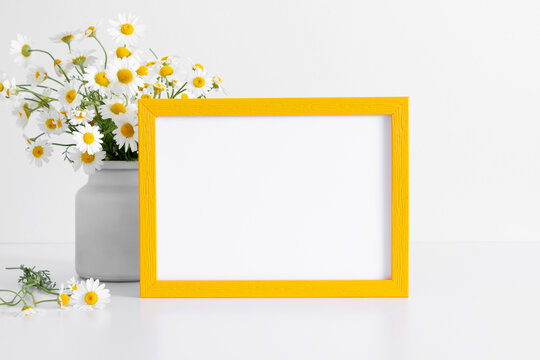 White chamomile flowers in vase and yellow photo frame on white table.