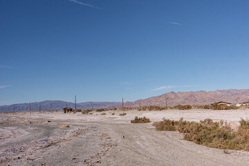 USA, CA, Salton Sea - December 28, 2012: White-gray salty sand of SE shoreline near Niland Marina with residential housing in back under blue sky and brown-green shrub in front.