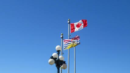 Canadian maple leaf and British Columbia flag flowing in the air. Blue sky and the lamp in the...