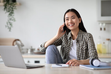 Smiling asian woman freelancer talking on phone and using laptop