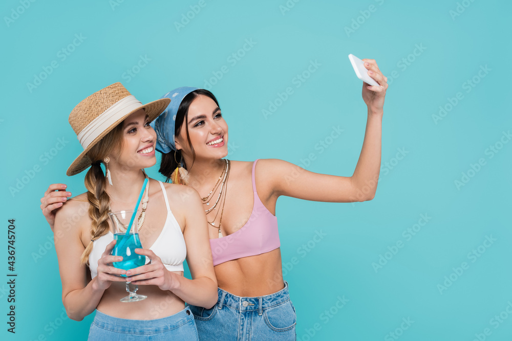 Wall mural smiling woman taking selfie with friend holding cocktail isolated on blue.