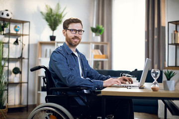 Side view of bearded young man sitting in wheelchair and typing on wireless laptop. Handsome disabled guy in eyewear working from home.