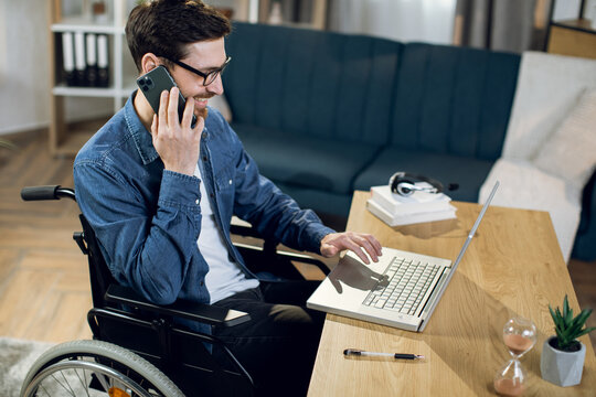 Disabled young man talking on smartphone and working on wireless laptop at home. Male freelancer sitting in wheelchair and using modern gadgets.