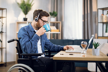 Side view of young man with special need using wireless laptop for remote work at home. Bearded guy sitting in wheelchair, drinking coffee and typing on computer.