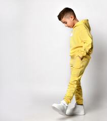 Fototapeta premium Profile view of a little brunette boy in yellow sportswear and white sneakers. Boy putting his hands posed posing on a white background. Concept of childhood, fashion and sports. Copy space.
