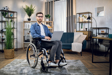Fototapeta na wymiar Young male freelancer sitting in wheelchair, using wireless laptop and looking at camera. Remote work for disabled people. Technology concept.