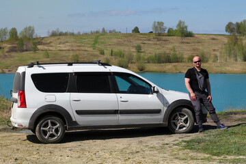 a man stands by a white car on the shore of a lake