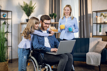 Fototapeta na wymiar Smiling woman holding two cups of coffee while standing near disabled husband that hugging cute daughter. Handicapped man working remotely on laptop to support family.